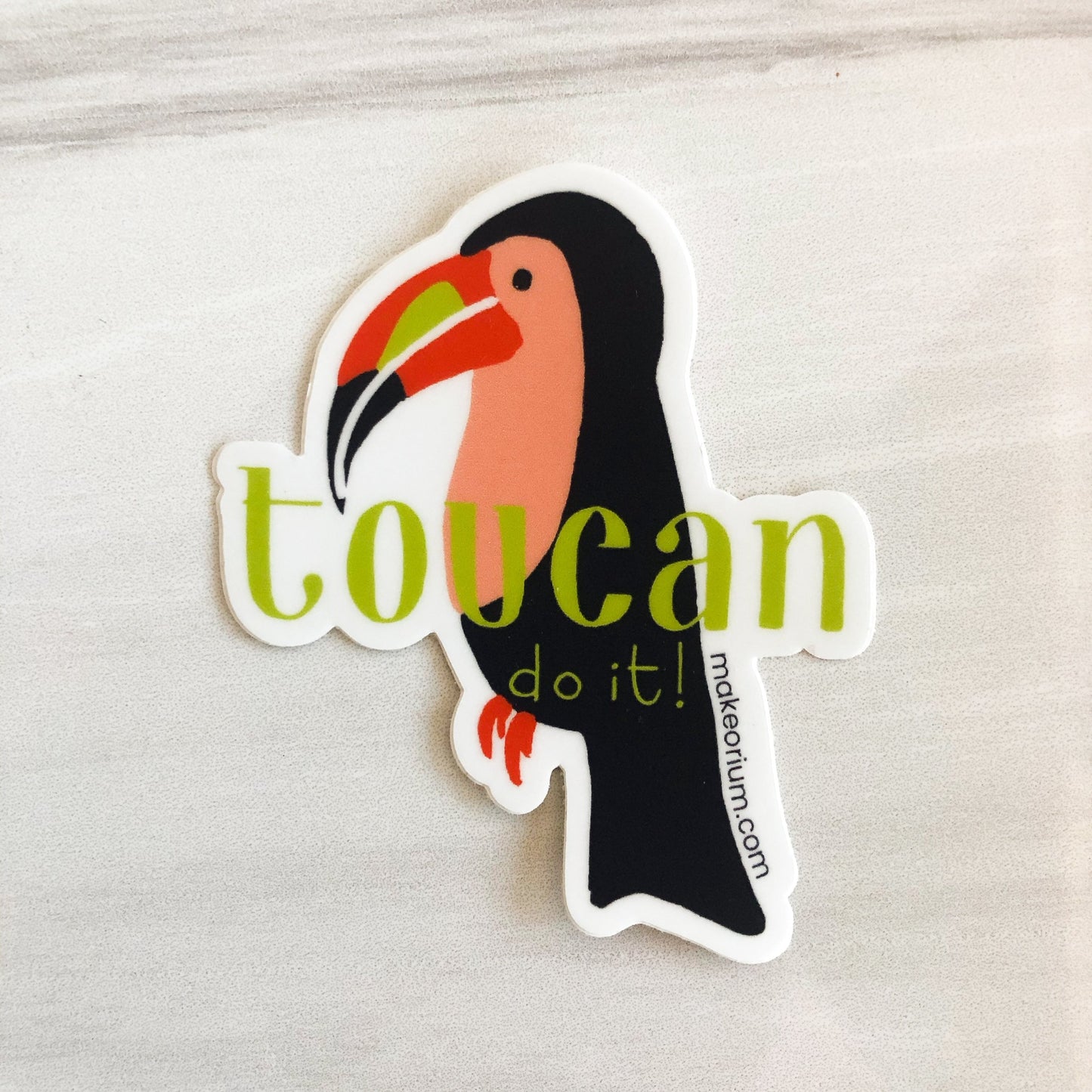 Vinyl sticker featuring an illustrated toucan in shades of blue, peach, orange, and lime green with hand-drawn text, "Toucan do it!" on a marble background.