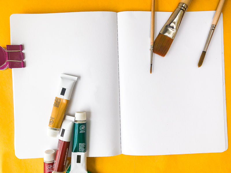 A blank sketchbook with paint tubes and paintbrushes