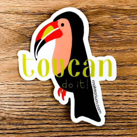 Vinyl sticker featuring an illustrated toucan with hand-drawn "Toucan do it!" text laying on a wood background.